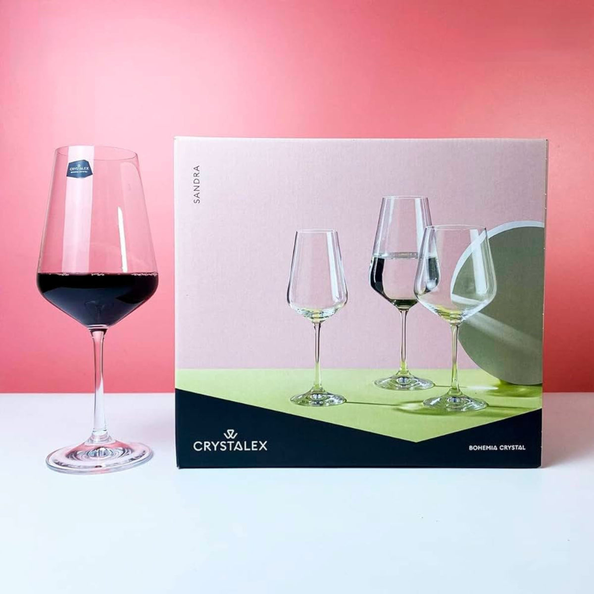 Wine glass with red wine next to a Crystalex packaging box on a pink background