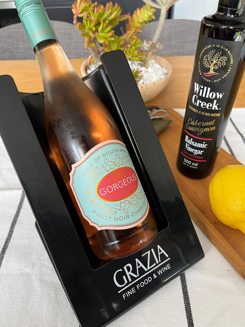 Bottle of wine with a decorative label in a black holder, accompanied by a bottle of Willow Creek balsamic vinegar on a wooden surface.